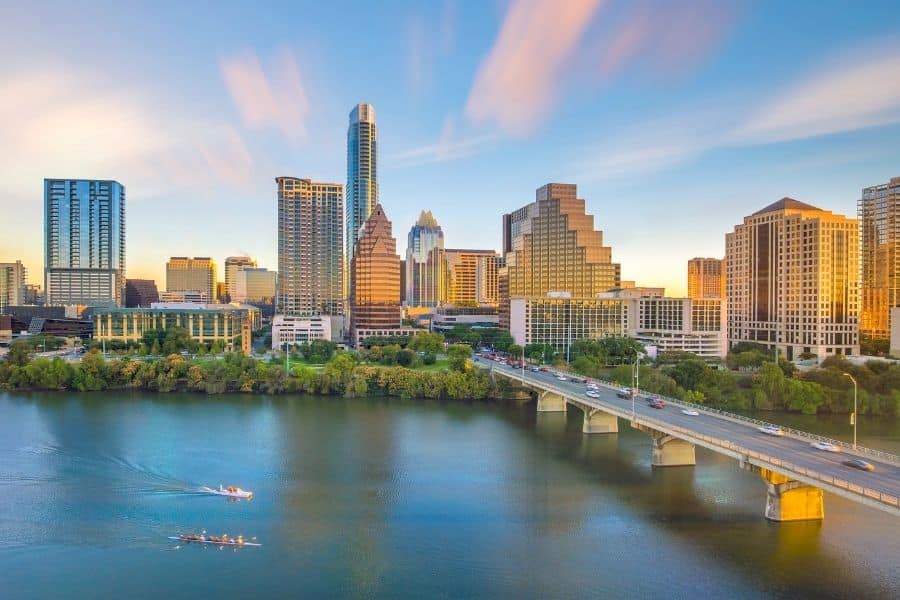 The Austin skyline at golden hour, viewed across the Colorado River with a bridge and rowing boats in the foreground