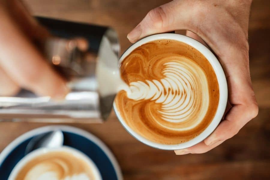 A barista's hands pouring steamed milk into an espresso to create a rosetta latte art pattern