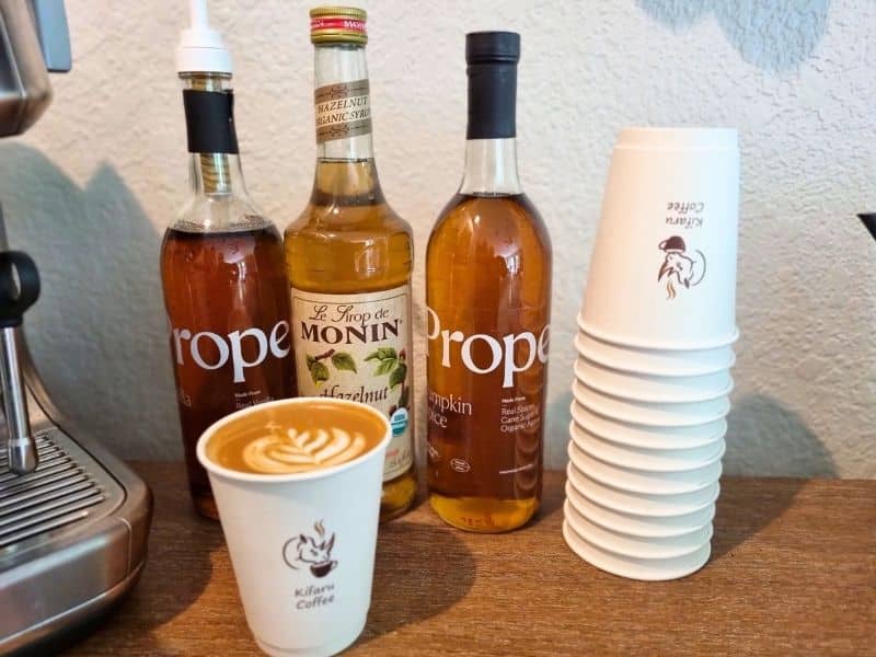 A Kifaru Coffee cup with latte art alongside Monin hazelnut and Proper organic flavored syrups, with a stack of branded cups
