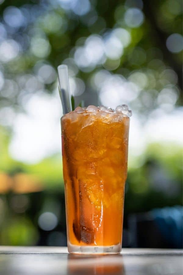 A tall glass of iced peach tea with a straw, served outdoors against a lush green background